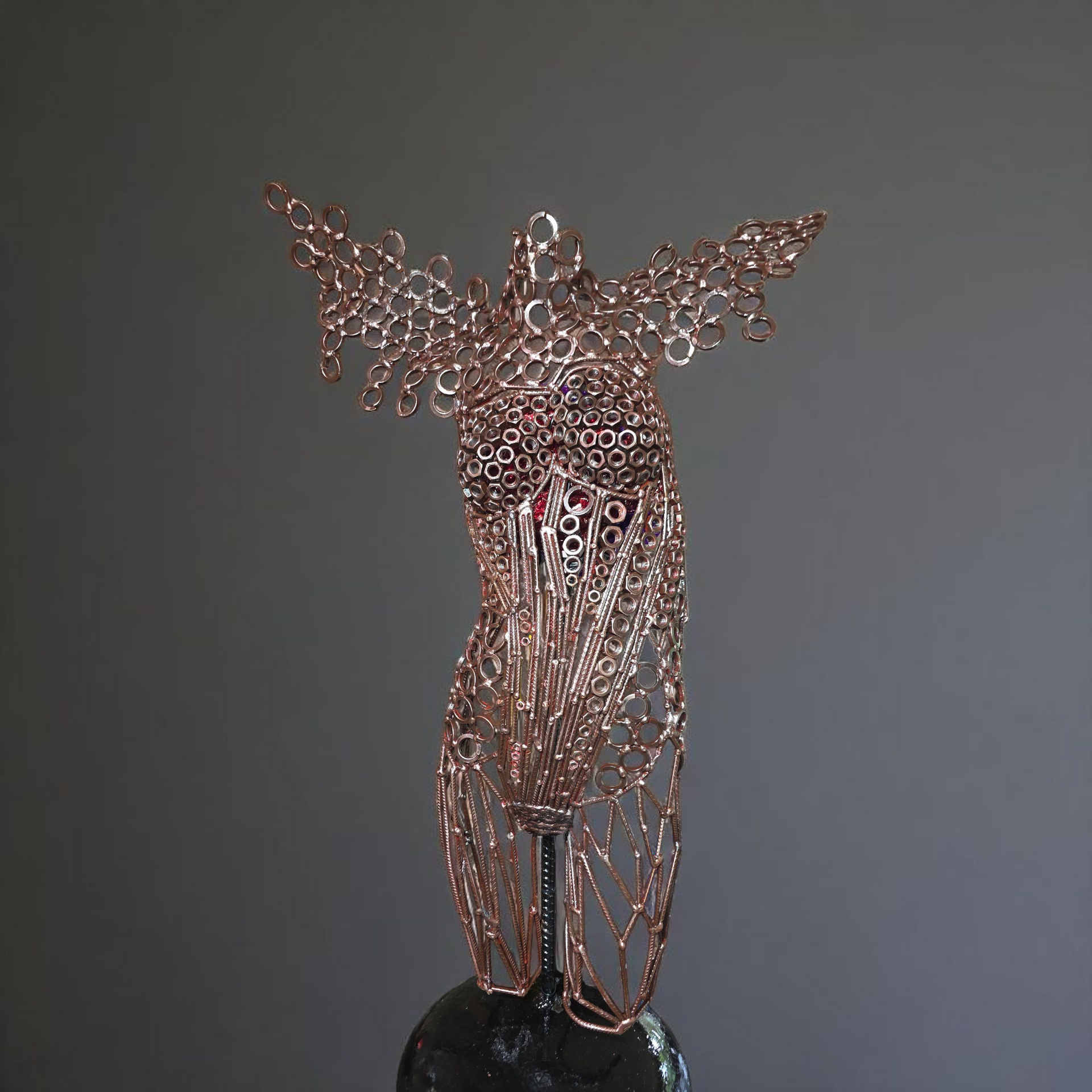 Wire sculpture of a human figure with outstretched wings, rose-colored wire, set on a dark base.