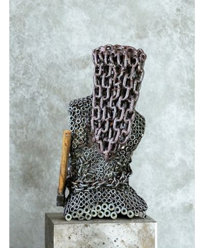 Metal sculpture of a headless figure draped in chains, holding a hammer; gray and rusty tones.