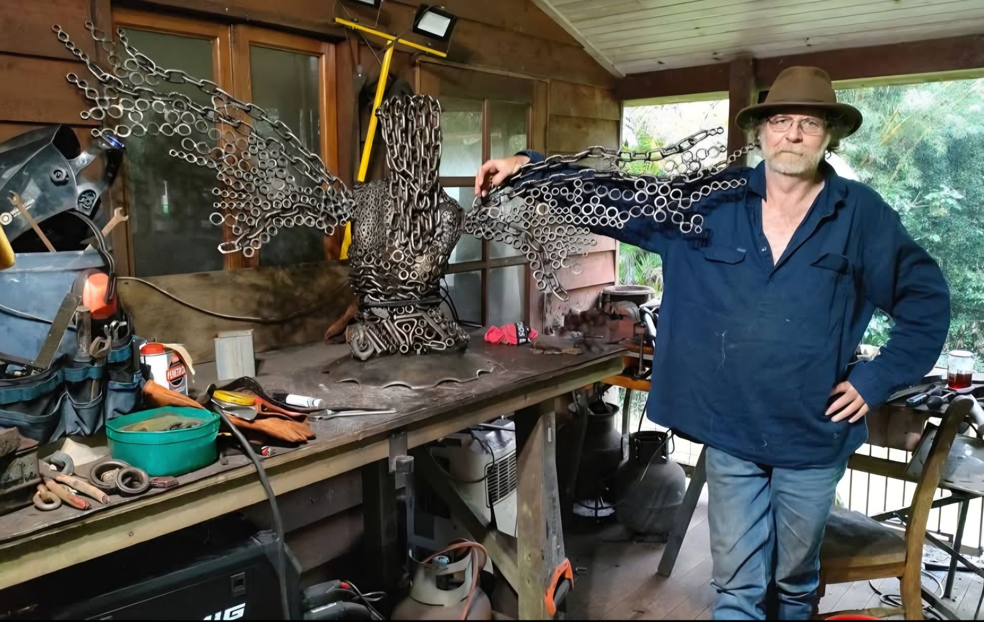 Man with metal eagle sculpture on workbench. He wears a hat, blue shirt, jeans, and stands on a porch.