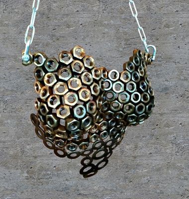 Metal sculpture of a bird, made of bolted nuts and hanging from a chain against a concrete wall.