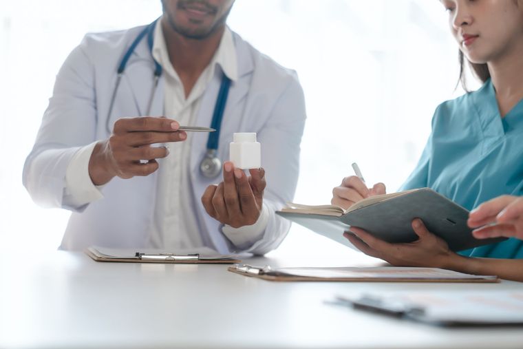 Doctor showing medication bottle to a person in scrubs writing notes at a table. Doctor showing medication bottle to a person in scrubs writing notes at a table.
