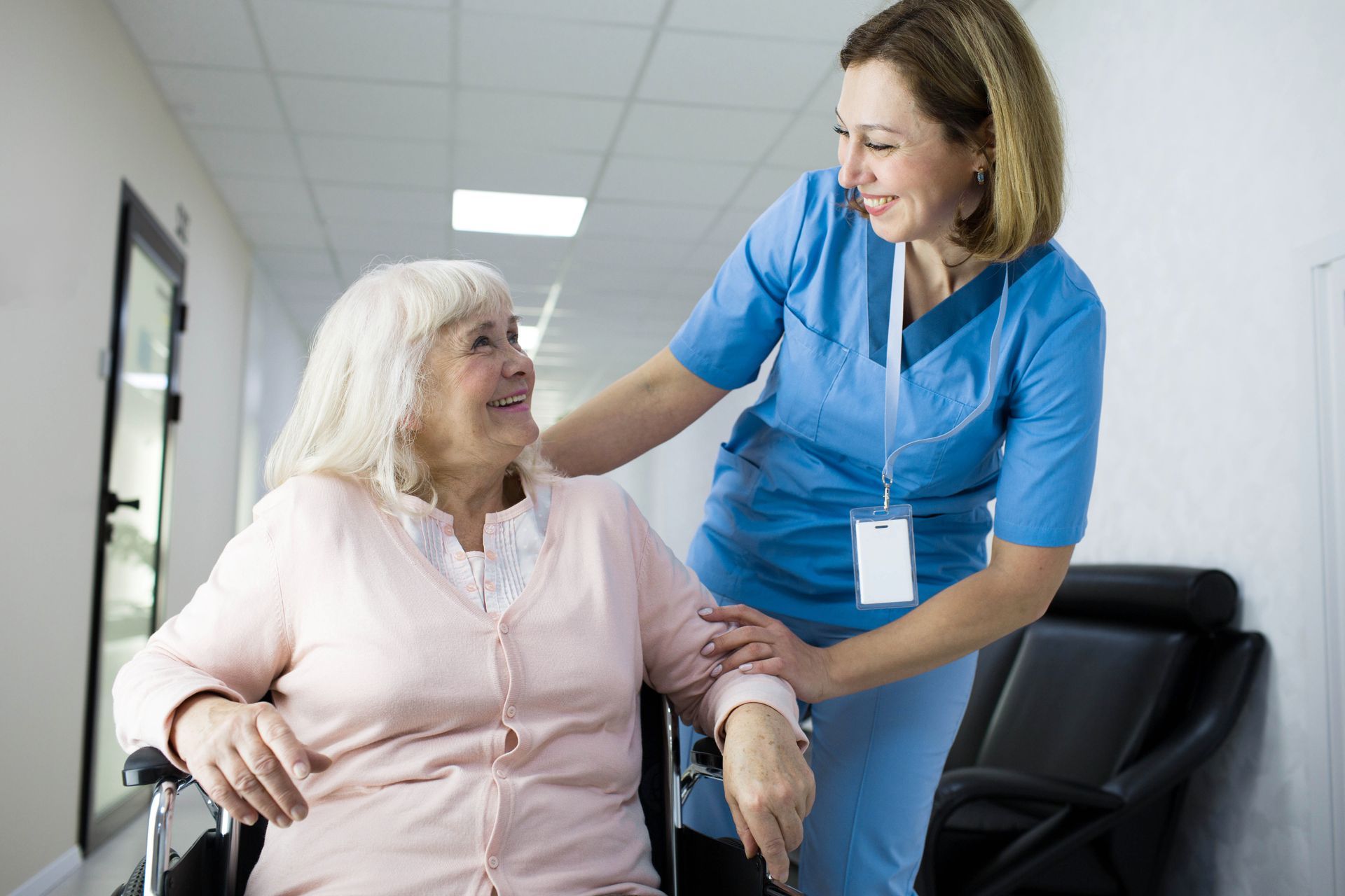 Nurse in blue scrubs assists elderly woman in wheelchair down a hallway.