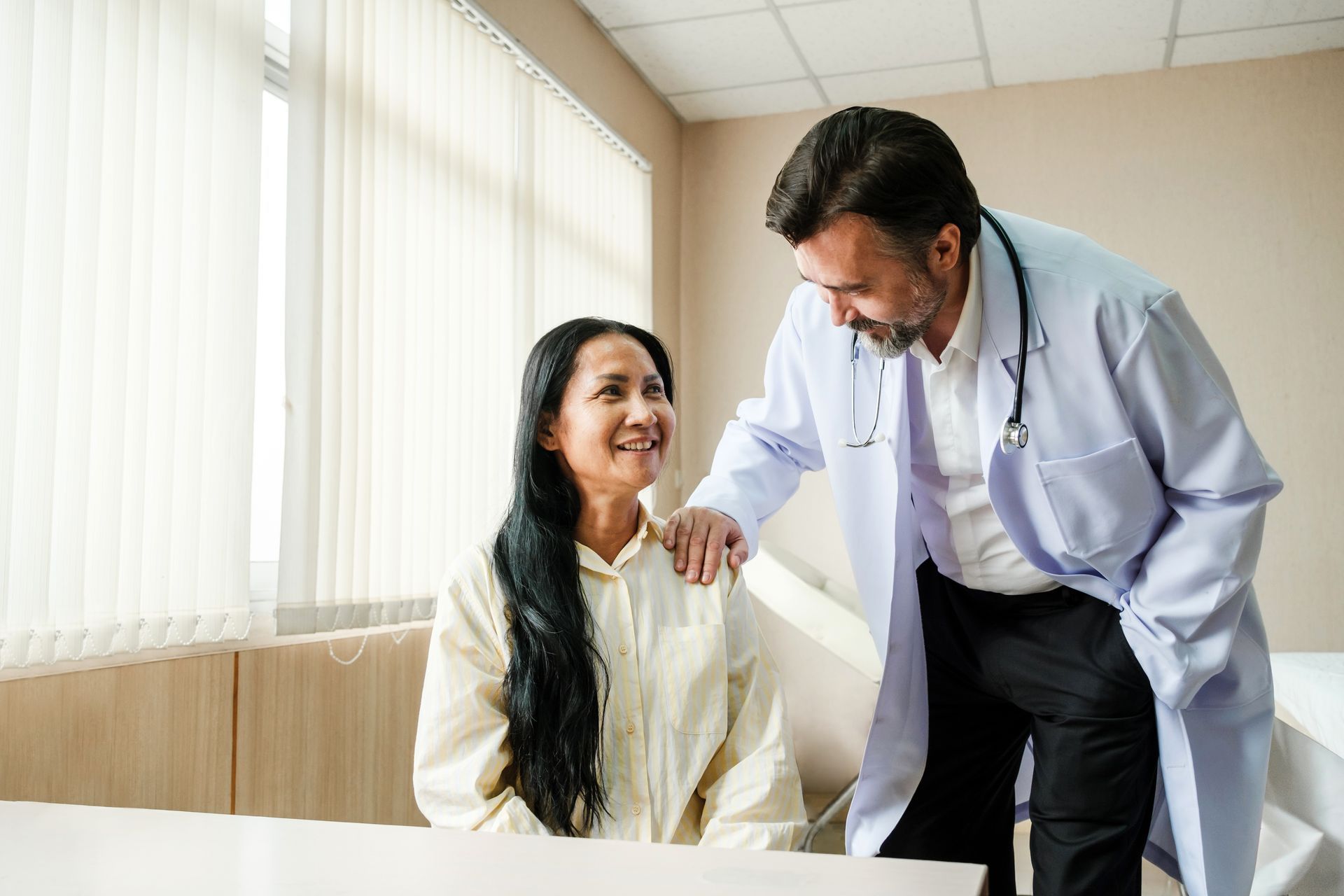 Doctor with stethoscope comforts a smiling patient in a brightly lit exam room.
