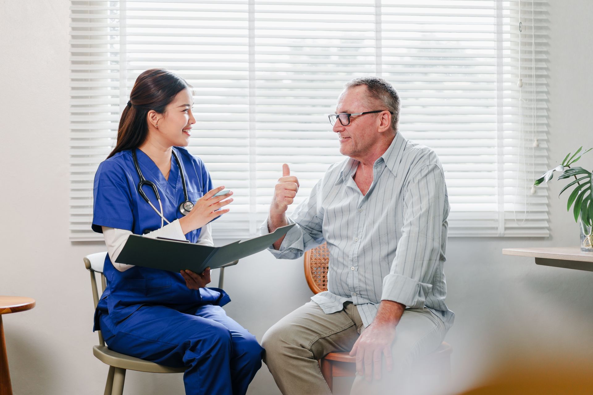 Nurse in blue scrubs consults with a patient in a well-lit room, patient giving a thumbs-up.
