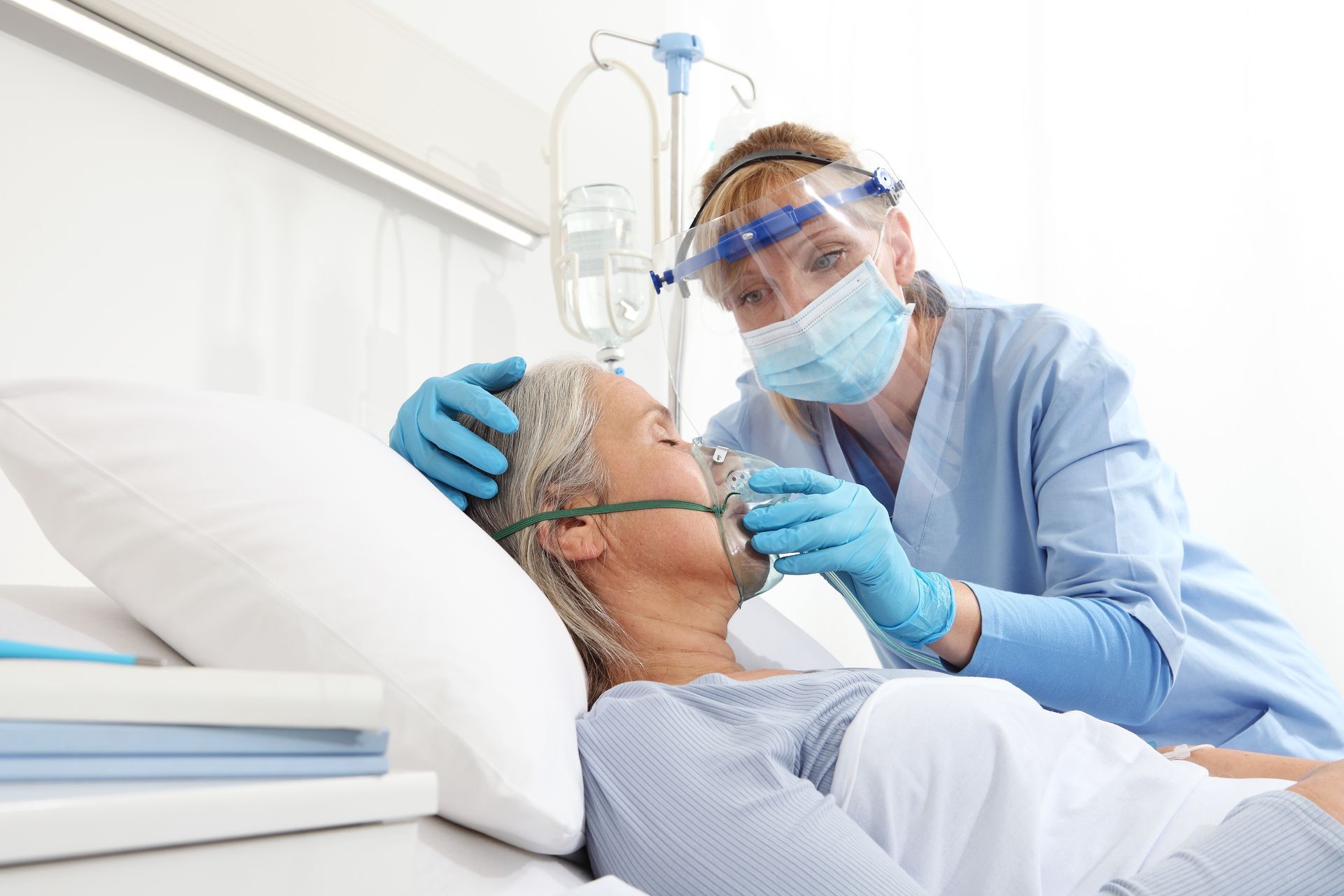 Nurse in blue scrubs, mask, and face shield assists a patient with an oxygen mask in a hospital bed.