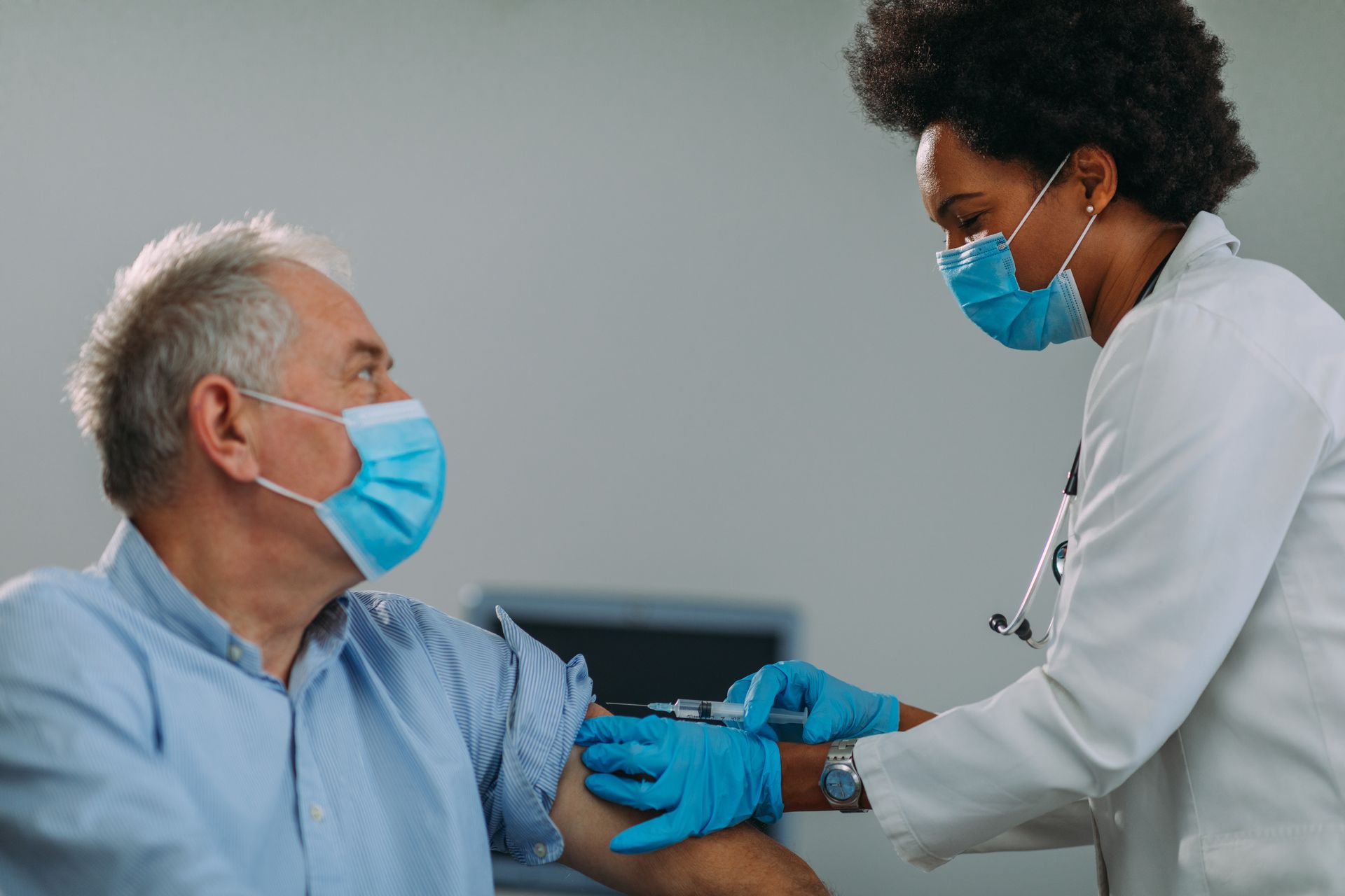 A masked medical professional giving an injection in the arm of a masked patient in a medical setting. A masked medical professional giving an injection in the arm of a masked patient in a medical setting.