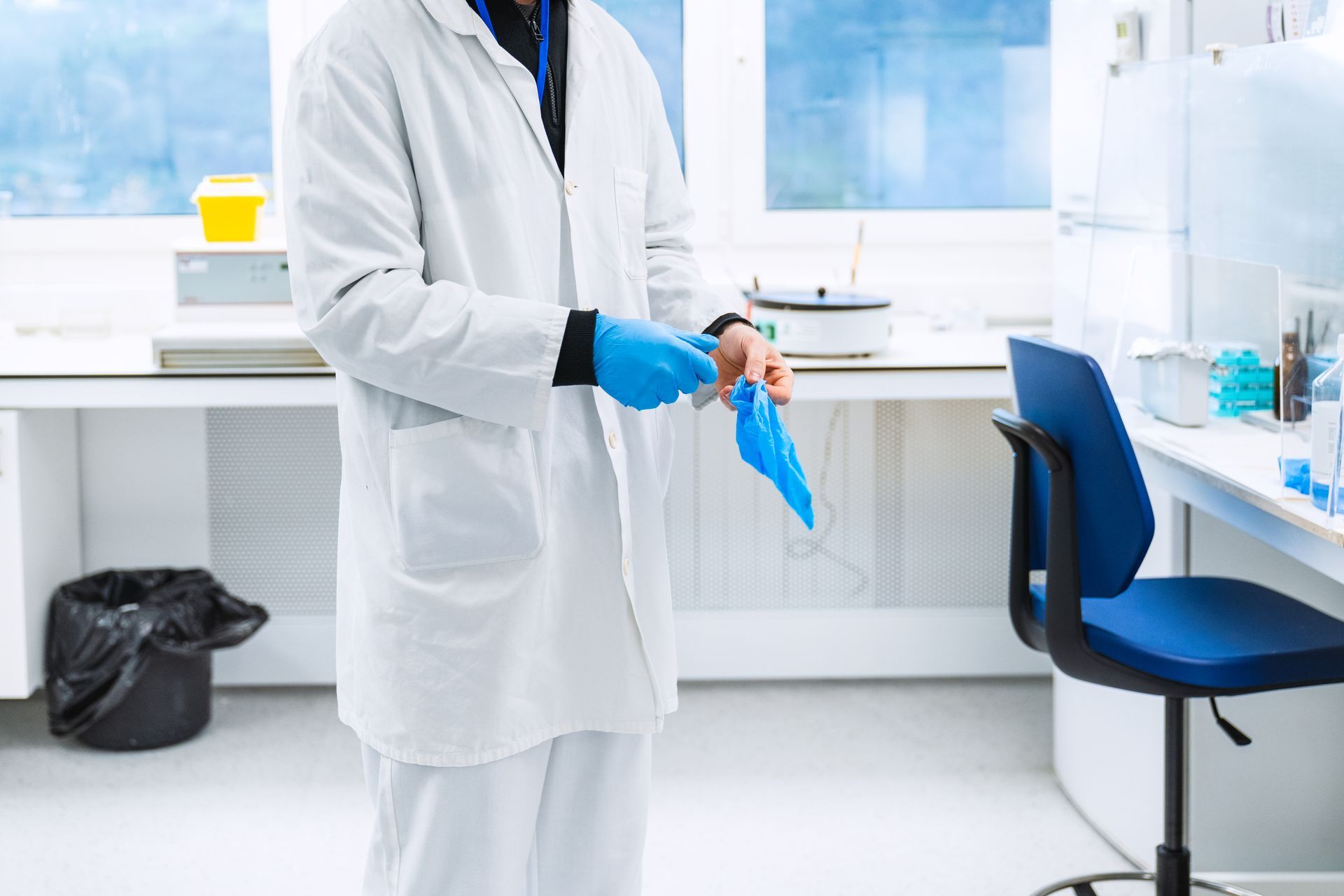 Person in a lab coat putting on blue gloves in a laboratory setting.