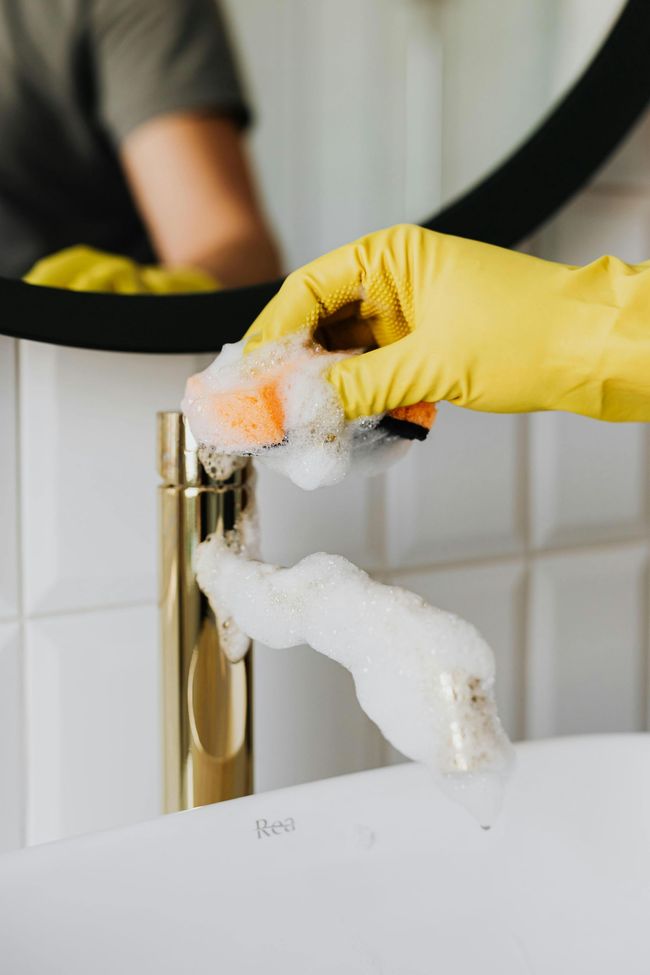 Yellow-gloved hand cleaning a sink faucet with a sponge and soap foam