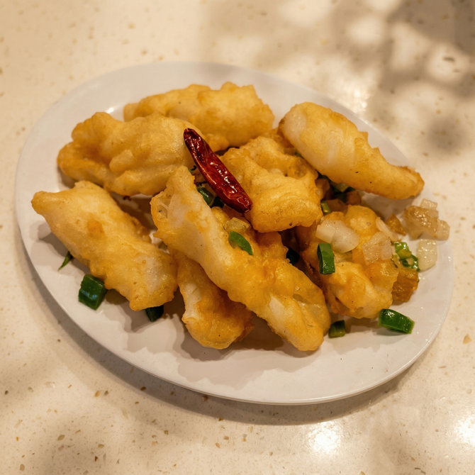 A white plate holds pieces of golden-brown fried calamari garnished with chopped green onions and a dried red chili.