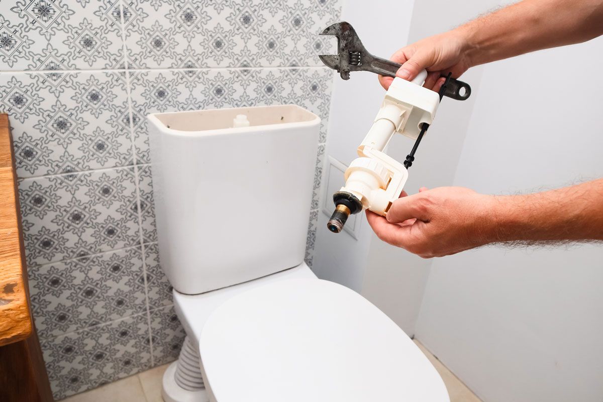 Person holding toilet fill valve and wrench near a toilet. Tile wall in background.