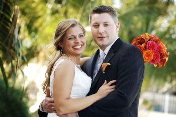 Couple in wedding attire smiling outdoors, with bright orange flowers in the background