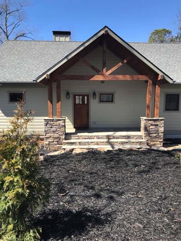 A light green house with a dark wood porch and stone pillars on a sunny day.