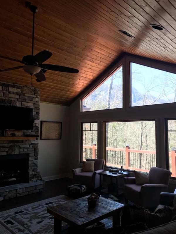 Cozy living room with stone fireplace, wood ceiling, large windows overlooking a deck and trees.