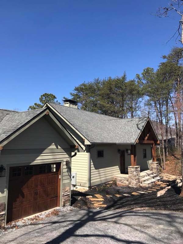 Green house with a brown garage door and stone steps, set in a wooded area under a blue sky.