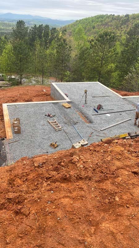 Construction site with foundation on gravel, surrounded by red soil, and trees in the background.