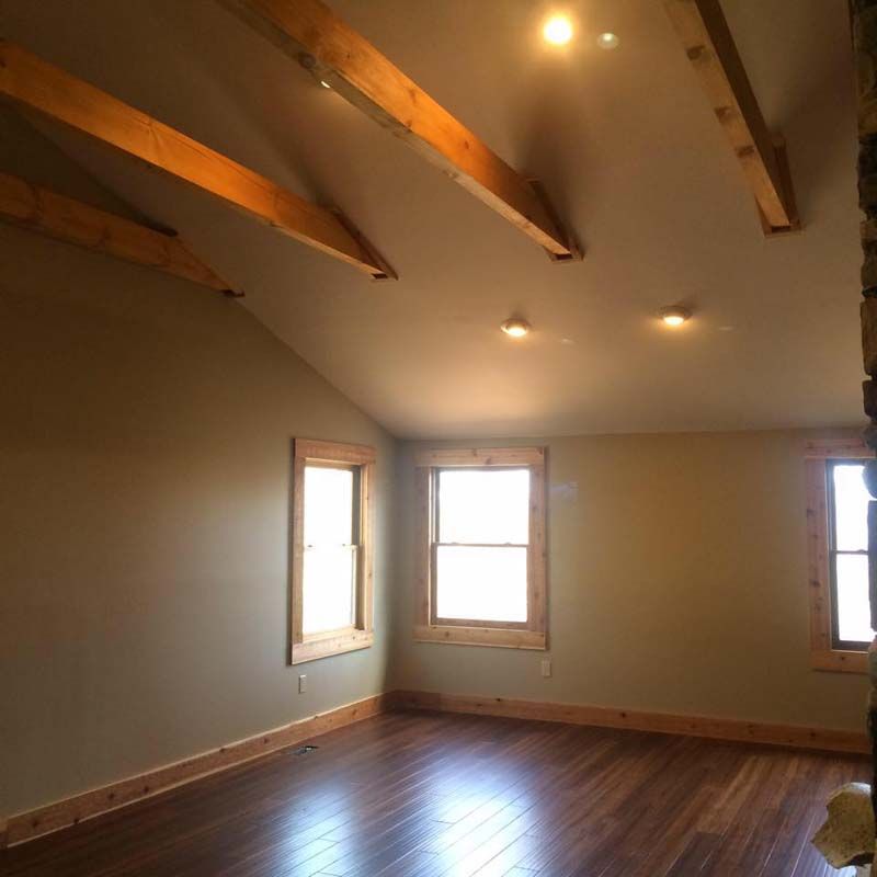Room with sloped ceiling, wooden beams, windows, hardwood floor, and light tan walls.