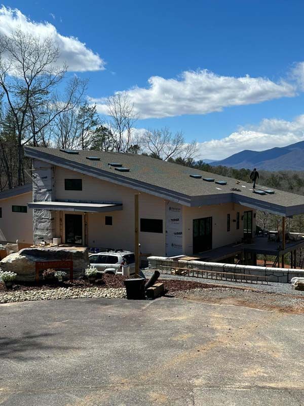 Modern house under construction with slanted roof, mountains in the background, blue sky with clouds.