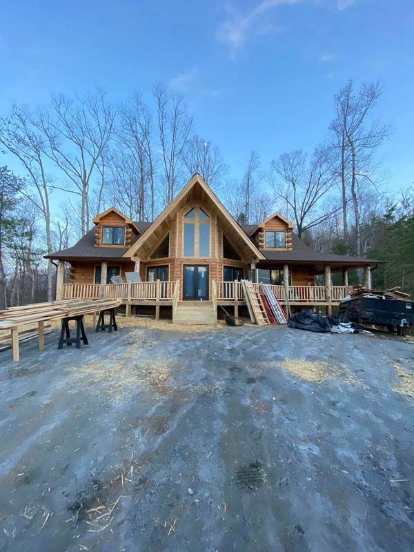 Log cabin under construction with a large front porch and trees in the background.