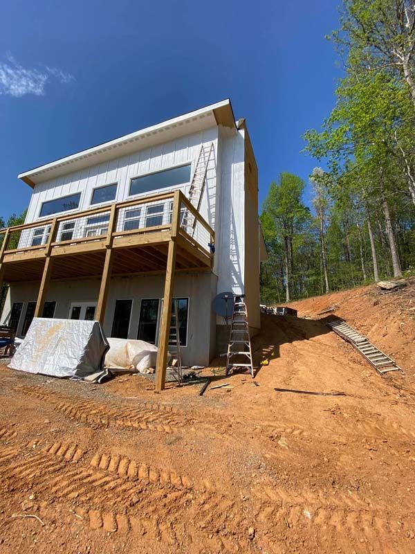 Two-story house with wooden deck under construction on a hillside. Exterior is white and tan, dirt ground.