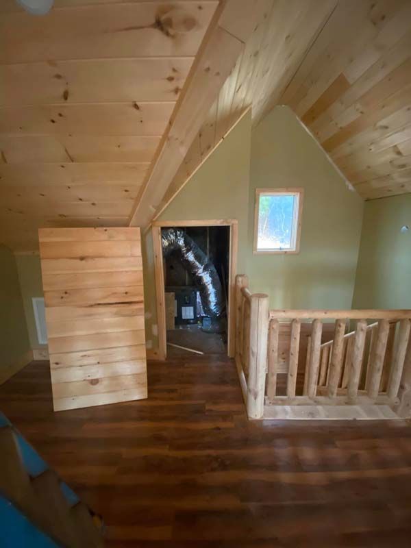 Wooden cabin interior with loft, wood flooring, log railing, and door to a dark room.