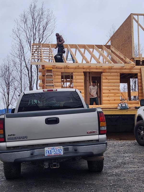 Construction of a log cabin; a worker on the roof, another inside. A truck is parked in front.