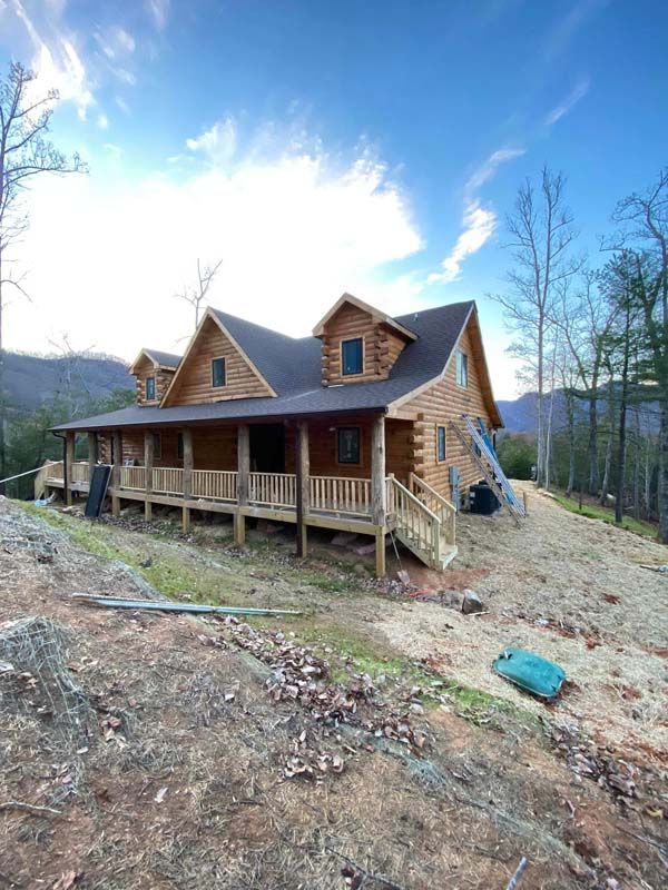 Log cabin with a wraparound porch and two dormers, set on a hillside with sparse vegetation.