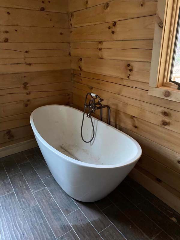 Freestanding white bathtub in a rustic bathroom with wood walls and gray tiled floor.