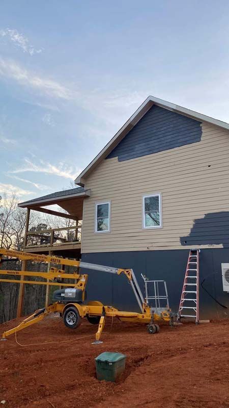 House exterior being painted, with a yellow lift machine and ladder on brown ground. Blue siding on upper section.