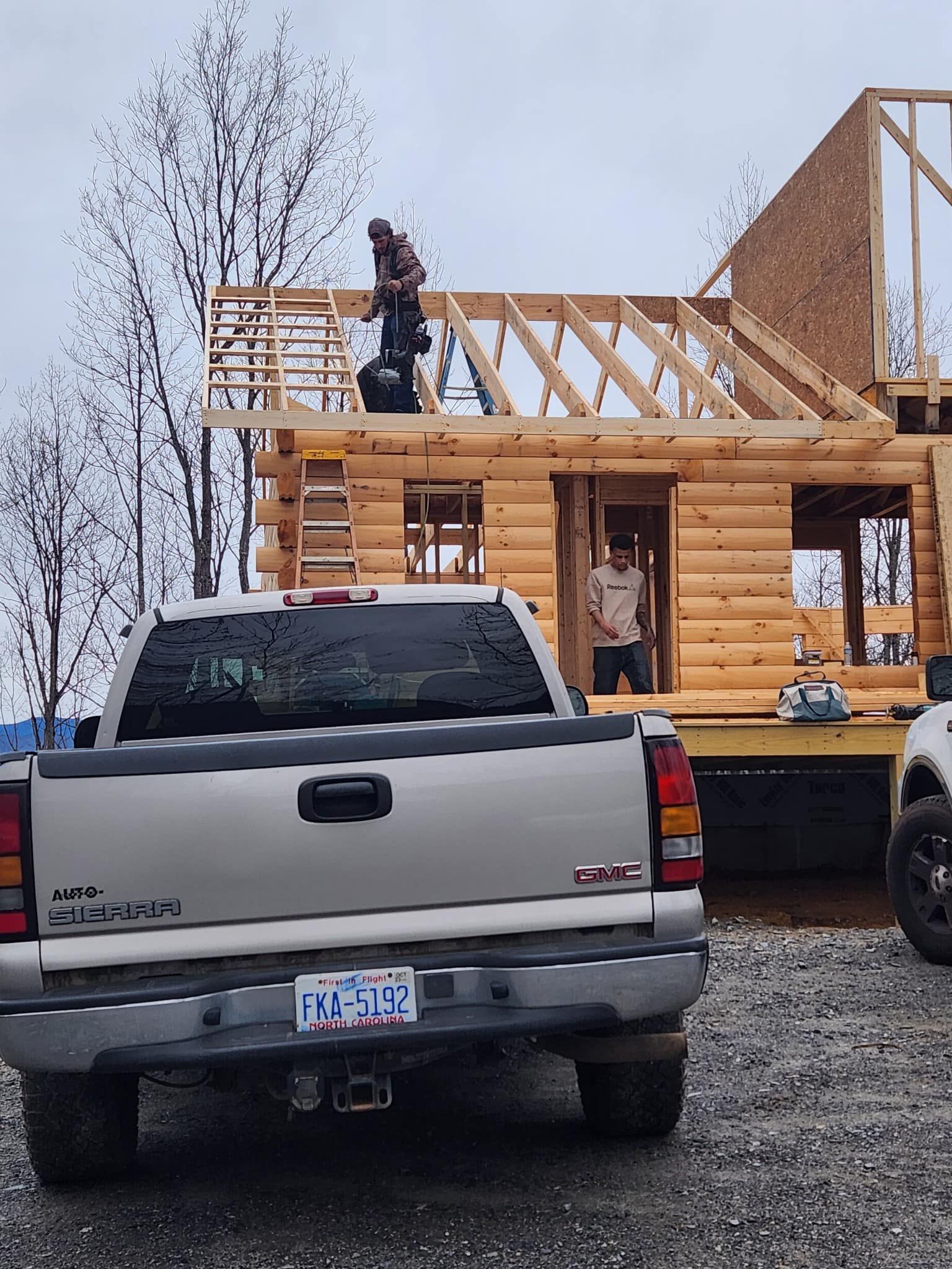 Construction workers building a log cabin, with a truck in the foreground and overcast sky.