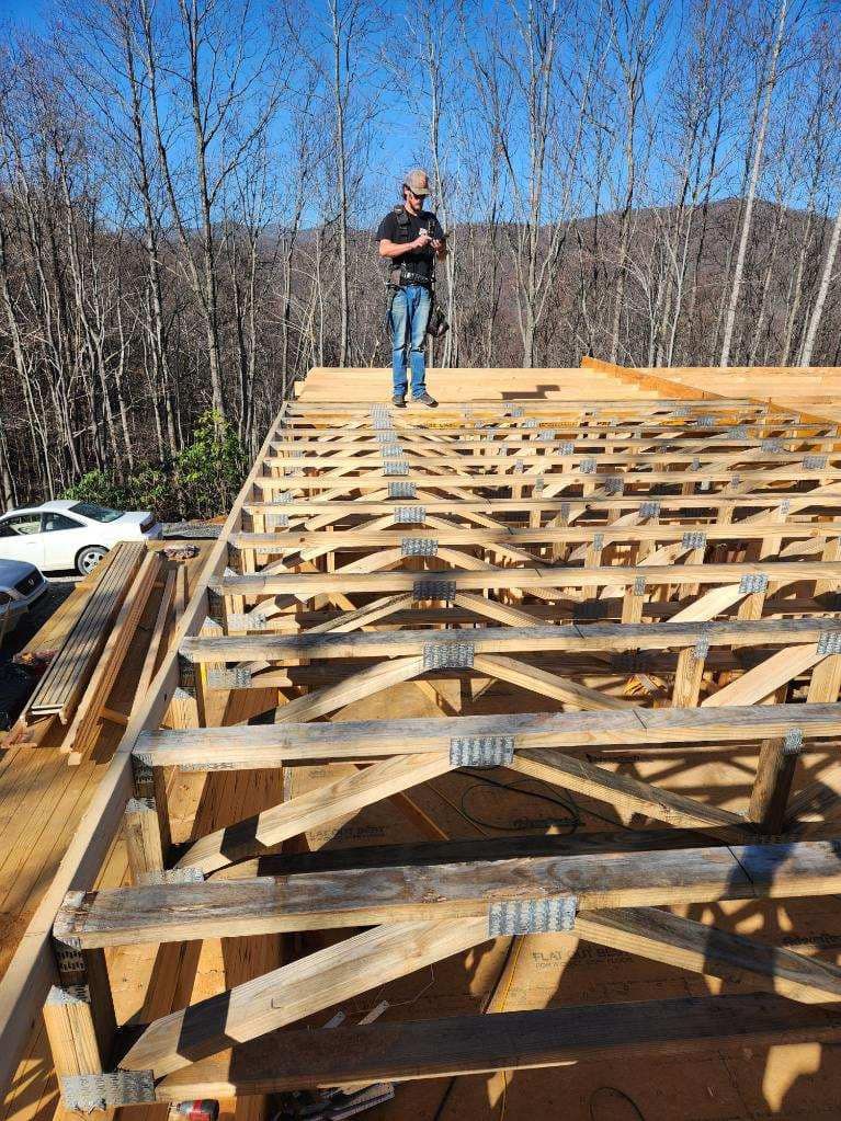 Man standing on wooden roof frame under construction, clear sunny sky.