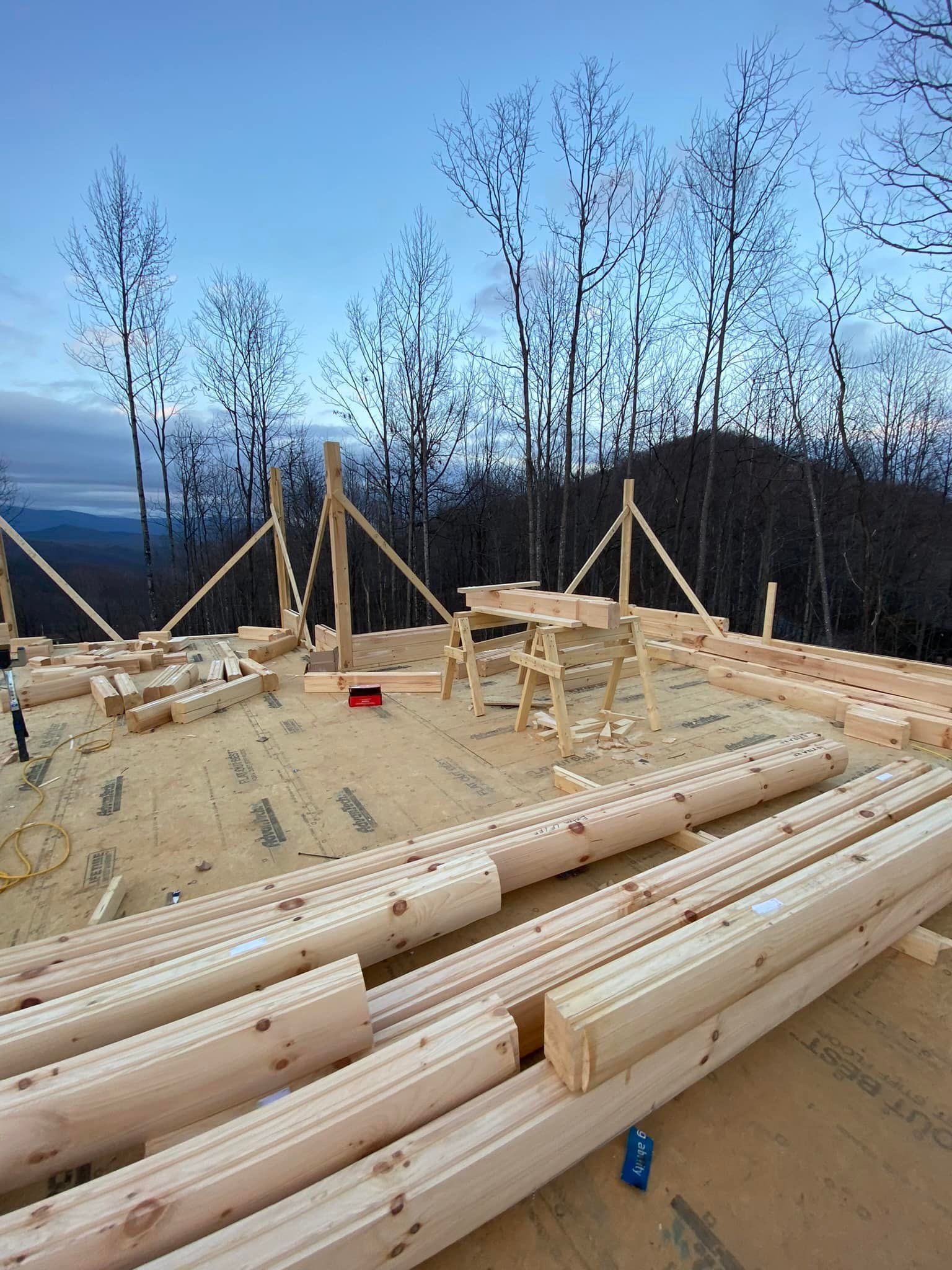 Construction site of a building, with logs, lumber, sawhorses, and tools visible on a wood platform surrounded by trees.