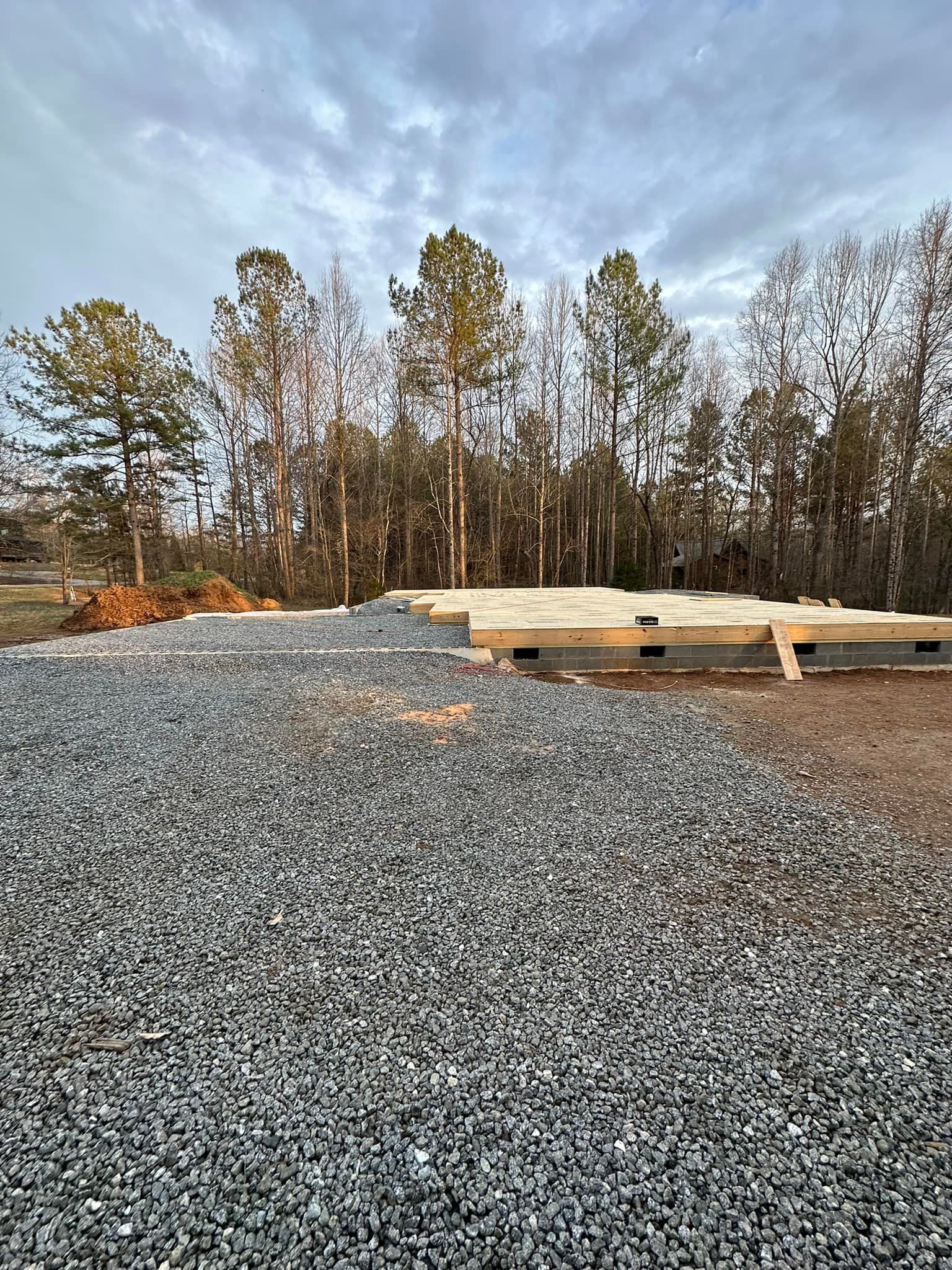 Construction site with gravel, wooden foundation, and trees under a cloudy sky.