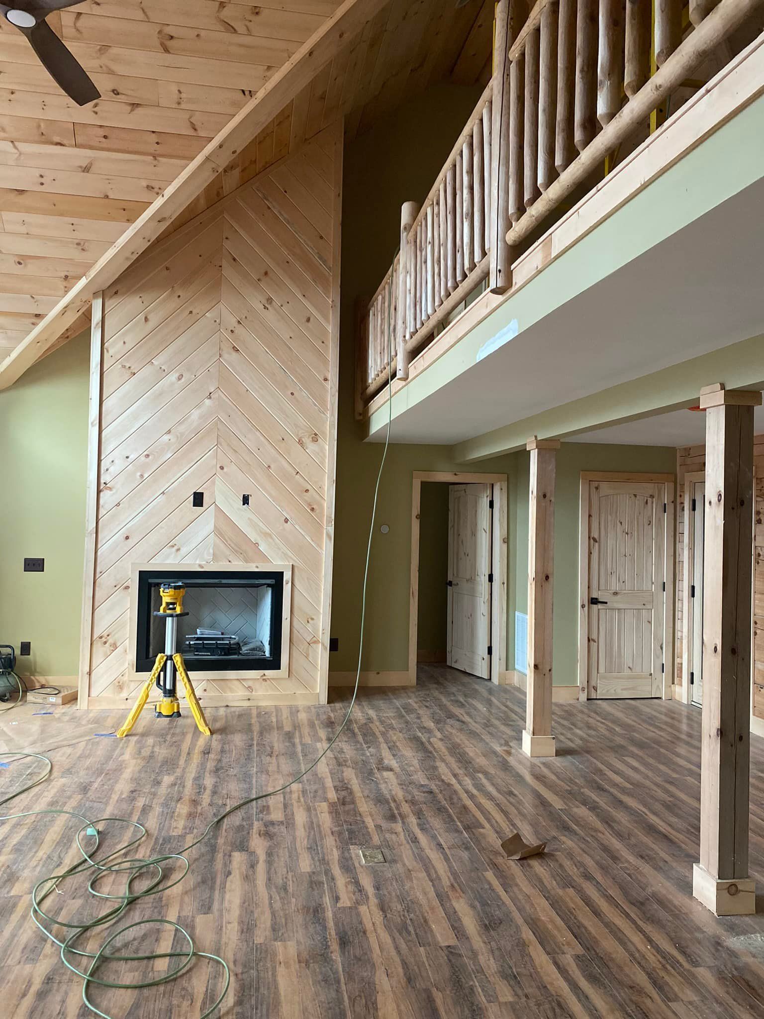 Interior of a cabin with fireplace and loft. Wood floors, light green walls, and natural wood beams are visible.