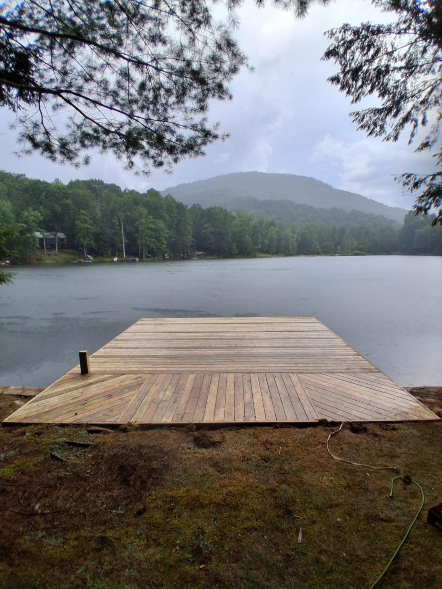 Wooden dock extending into a lake, surrounded by trees and a distant mountain, under a cloudy sky.