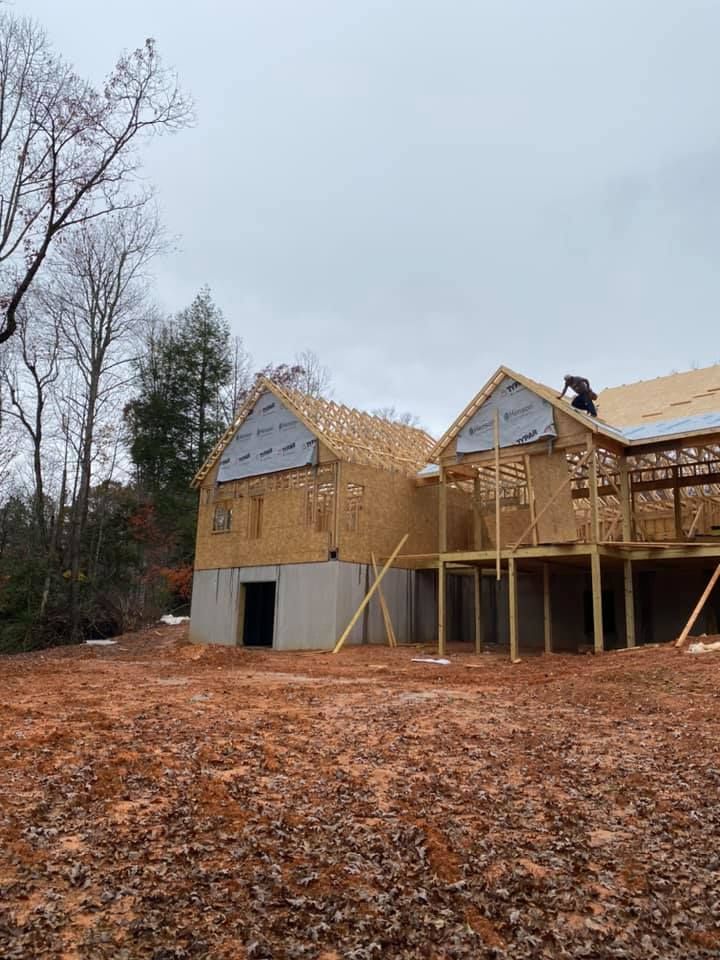 Wood-framed house under construction; a worker on the roof, cloudy day, brown dirt, trees in the background.