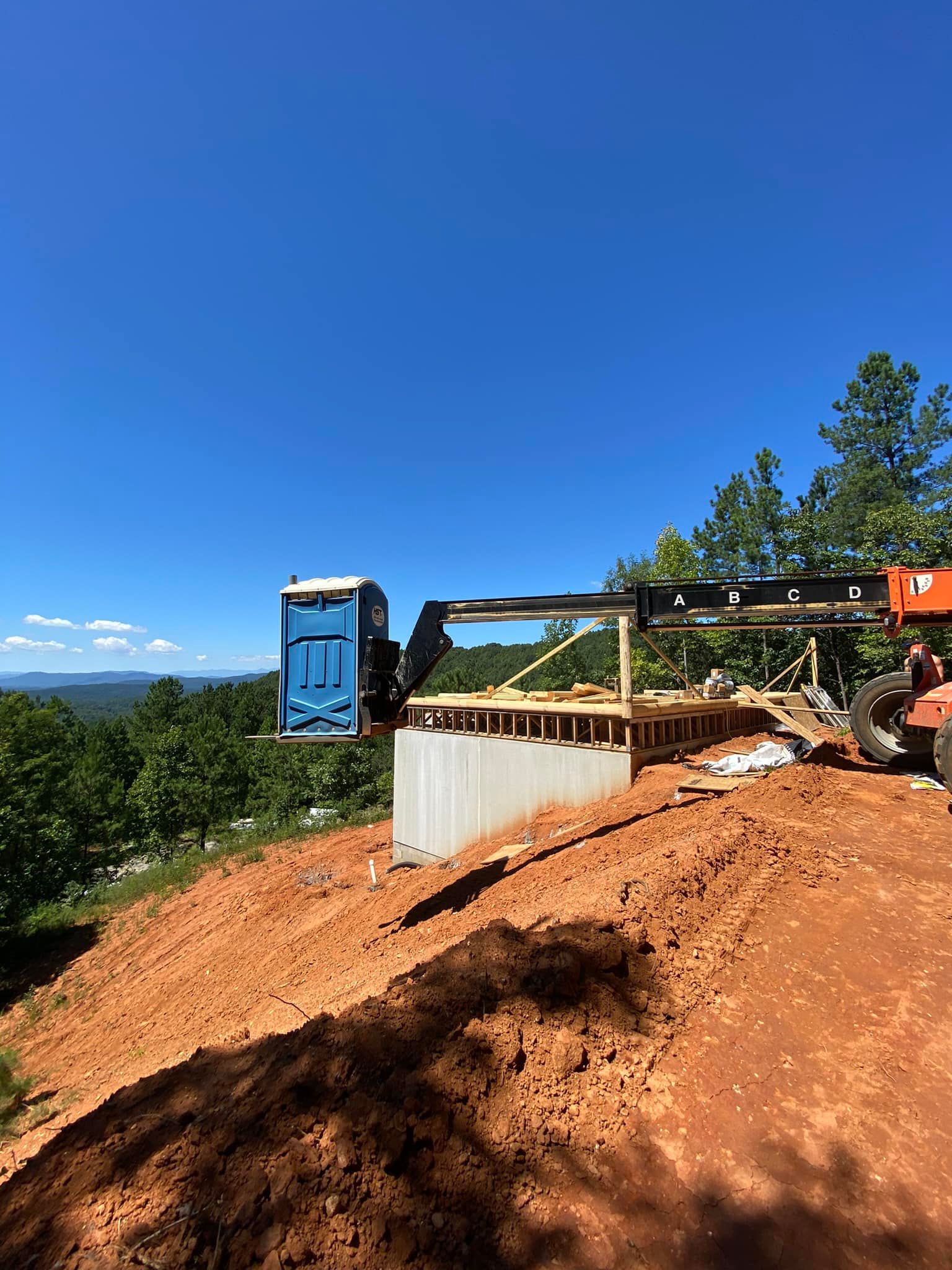 A forklift lifting a portable toilet to a construction site overlooking a landscape.