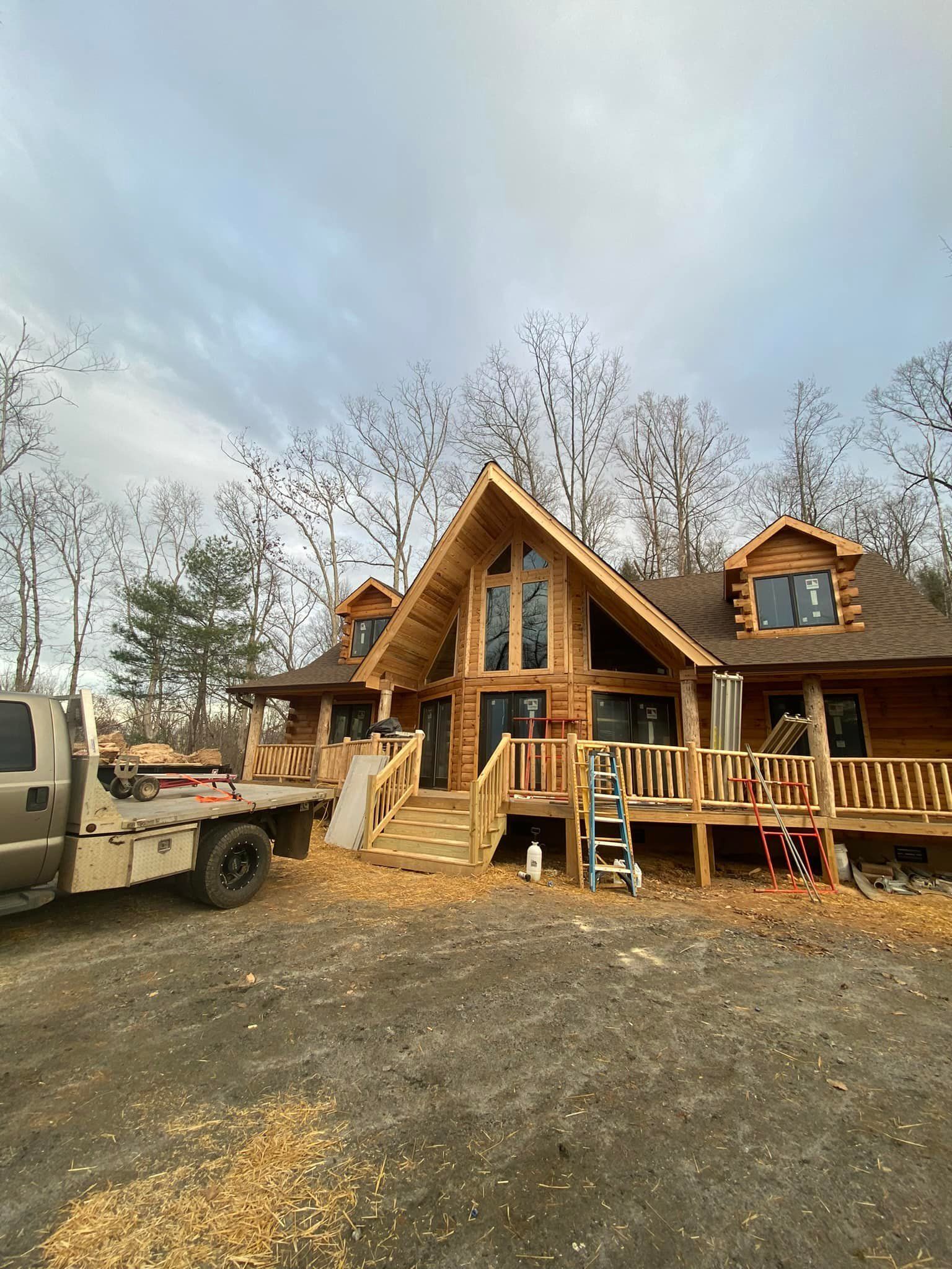 Log cabin under construction with a deck, large windows, and a truck parked outside.
