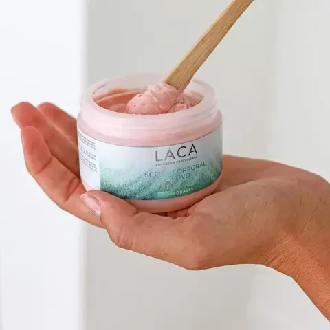 Una mano sostiene un frasco abierto de exfoliante corporal rosa con un aplicador de madera en su interior.