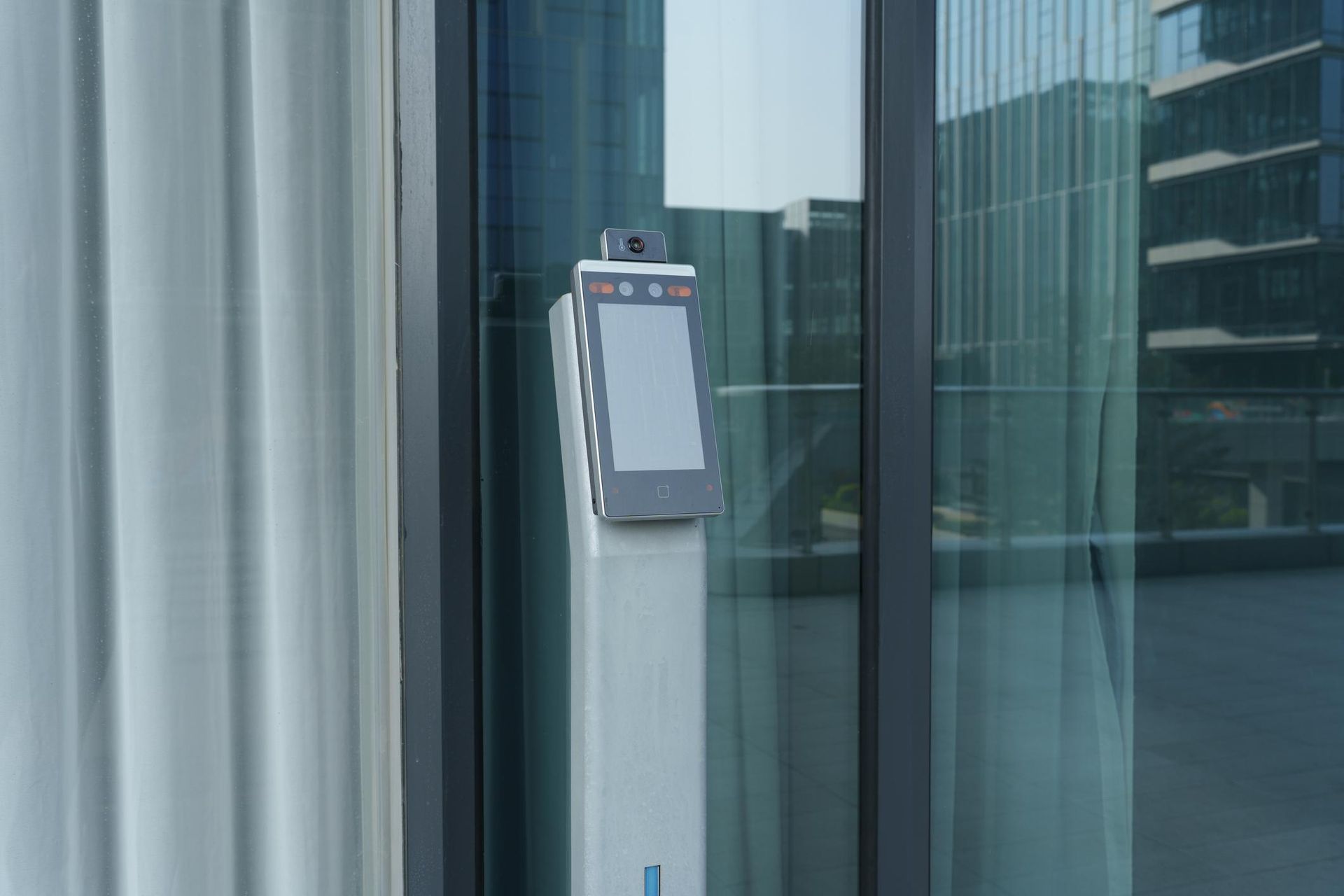 A standing biometric scanner with a blank digital screen positioned in front of a glass office building facade.