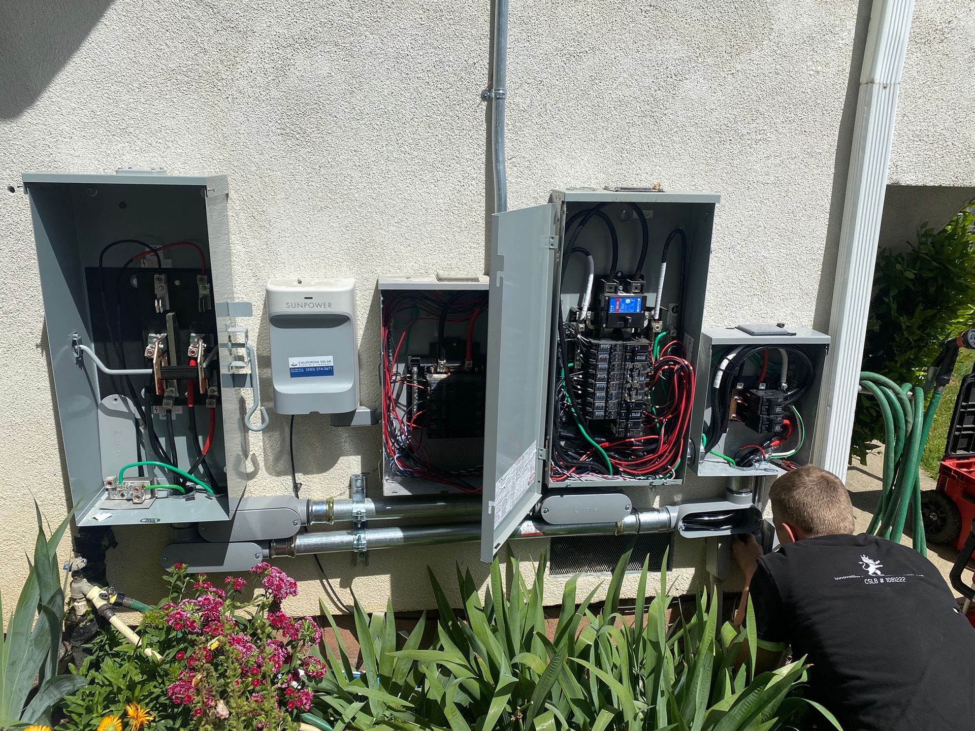 An electrician working on an outdoor wall-mounted electrical panel system with multiple open metal boxes and wiring.