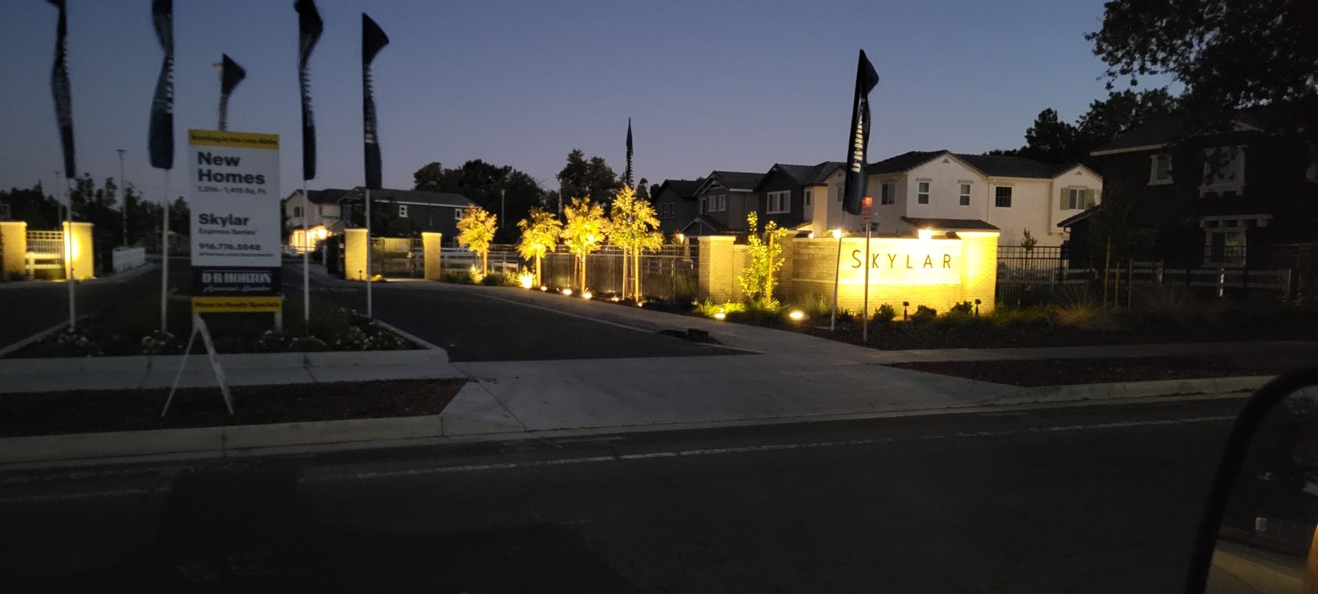 An illuminated entrance to a residential housing development at dusk, featuring landscaped trees and branded signage.