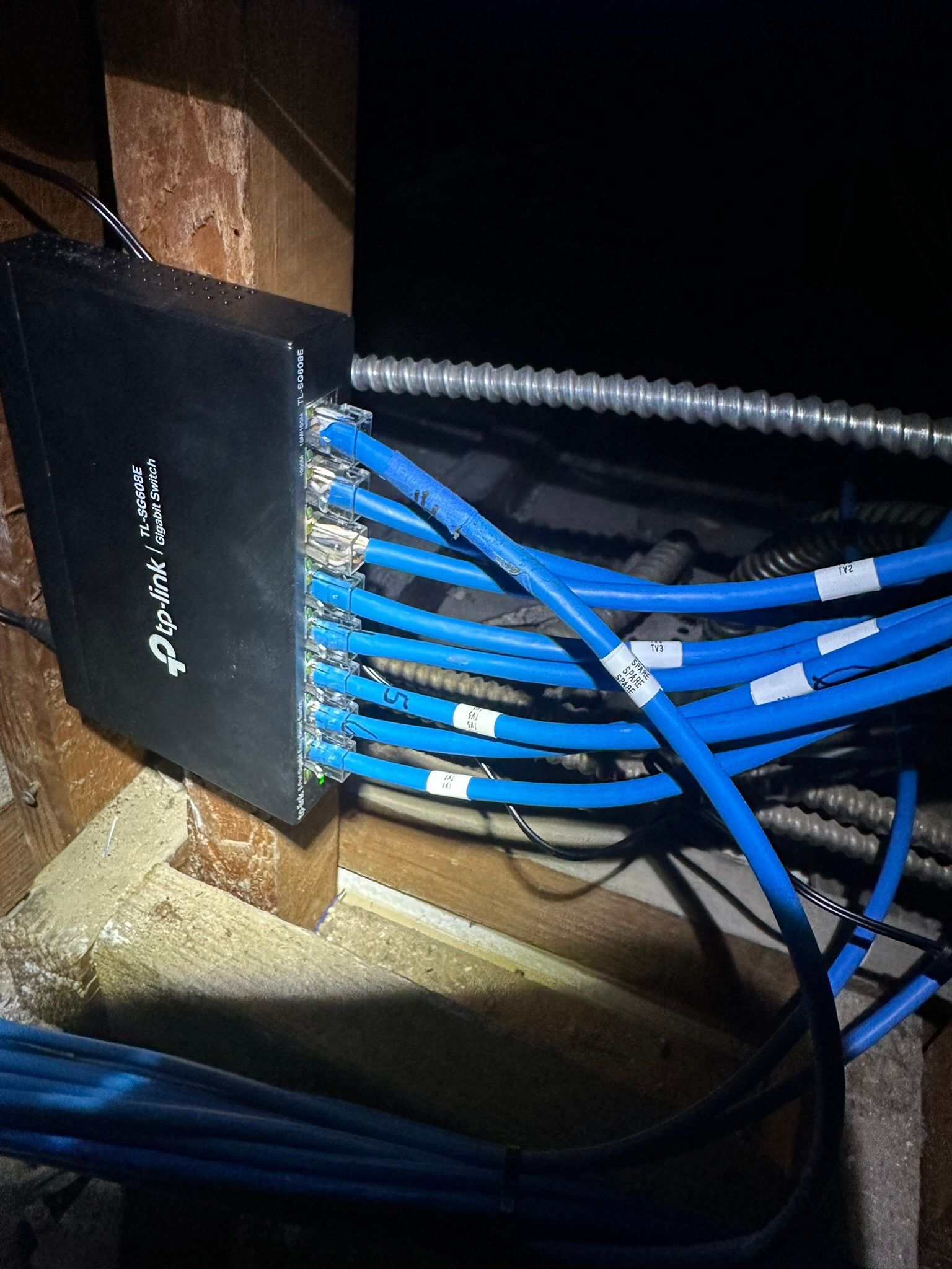 A black network switch mounted on a wooden beam in an attic, with several blue Ethernet cables plugged into its side.