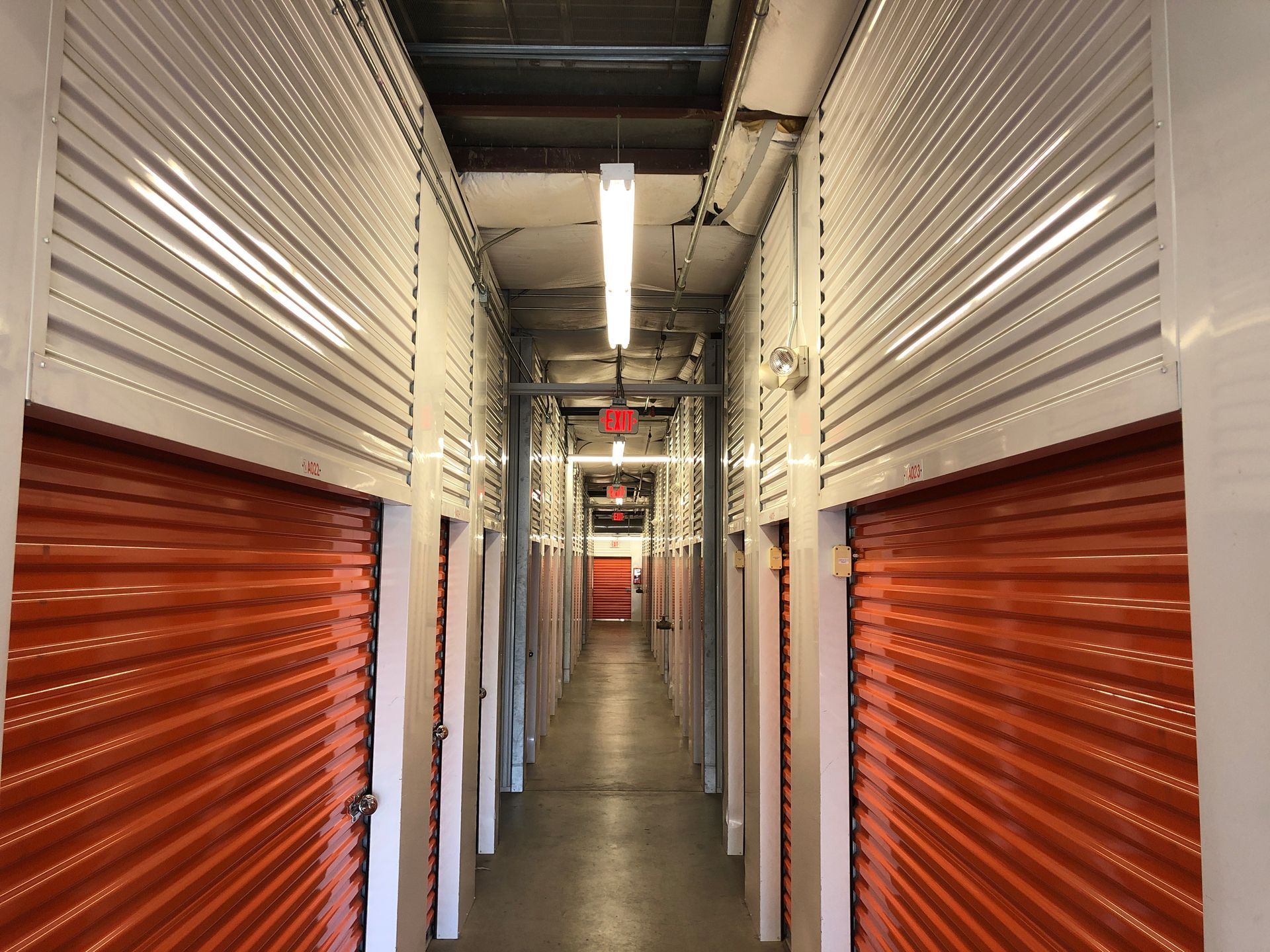 A hallway in a storage facility lined with rows of closed orange roll-up doors and white metal partitions.
