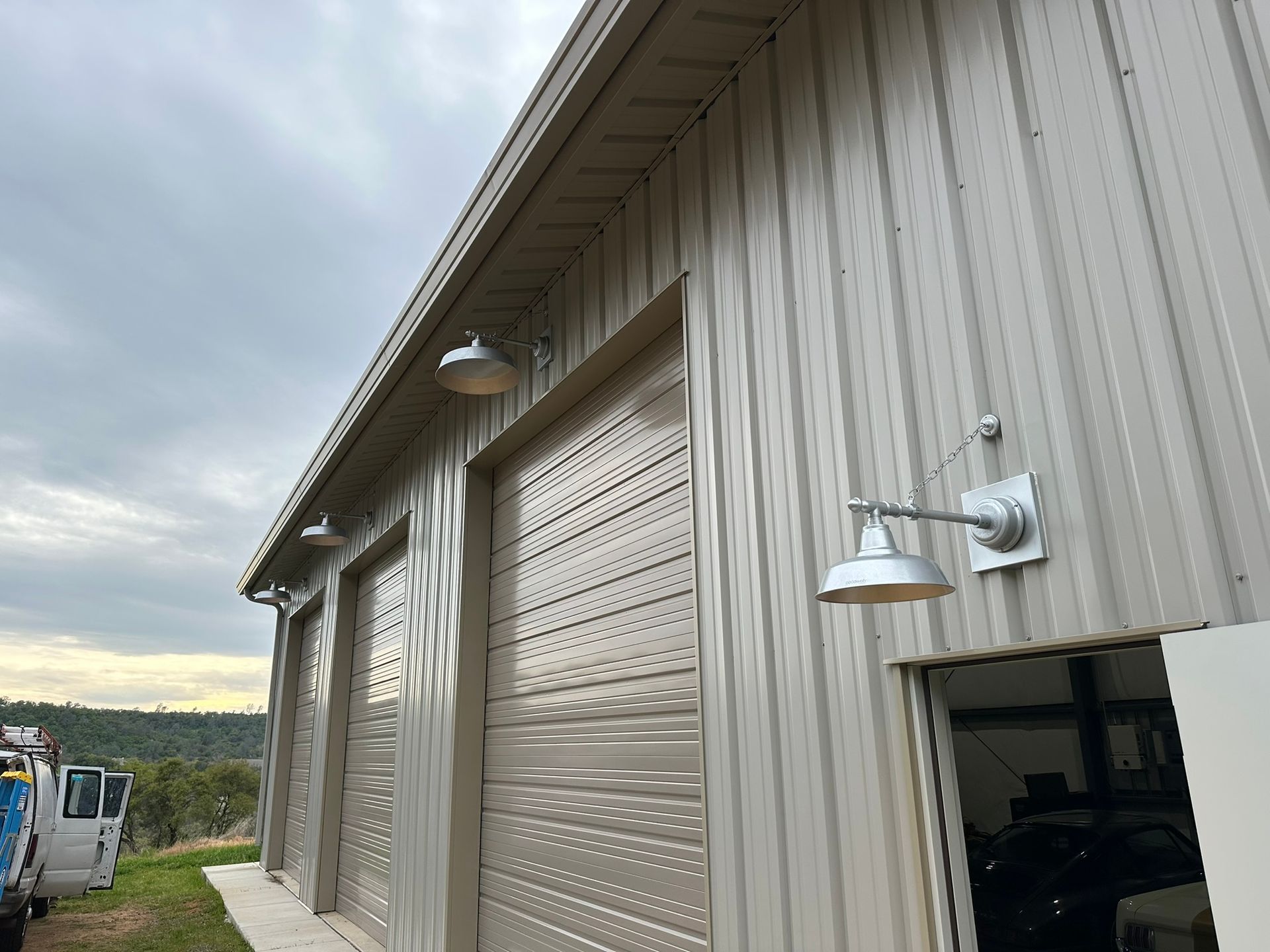 Light-colored metal building with multiple roll-up doors and gooseneck wall-mounted lights outdoors.