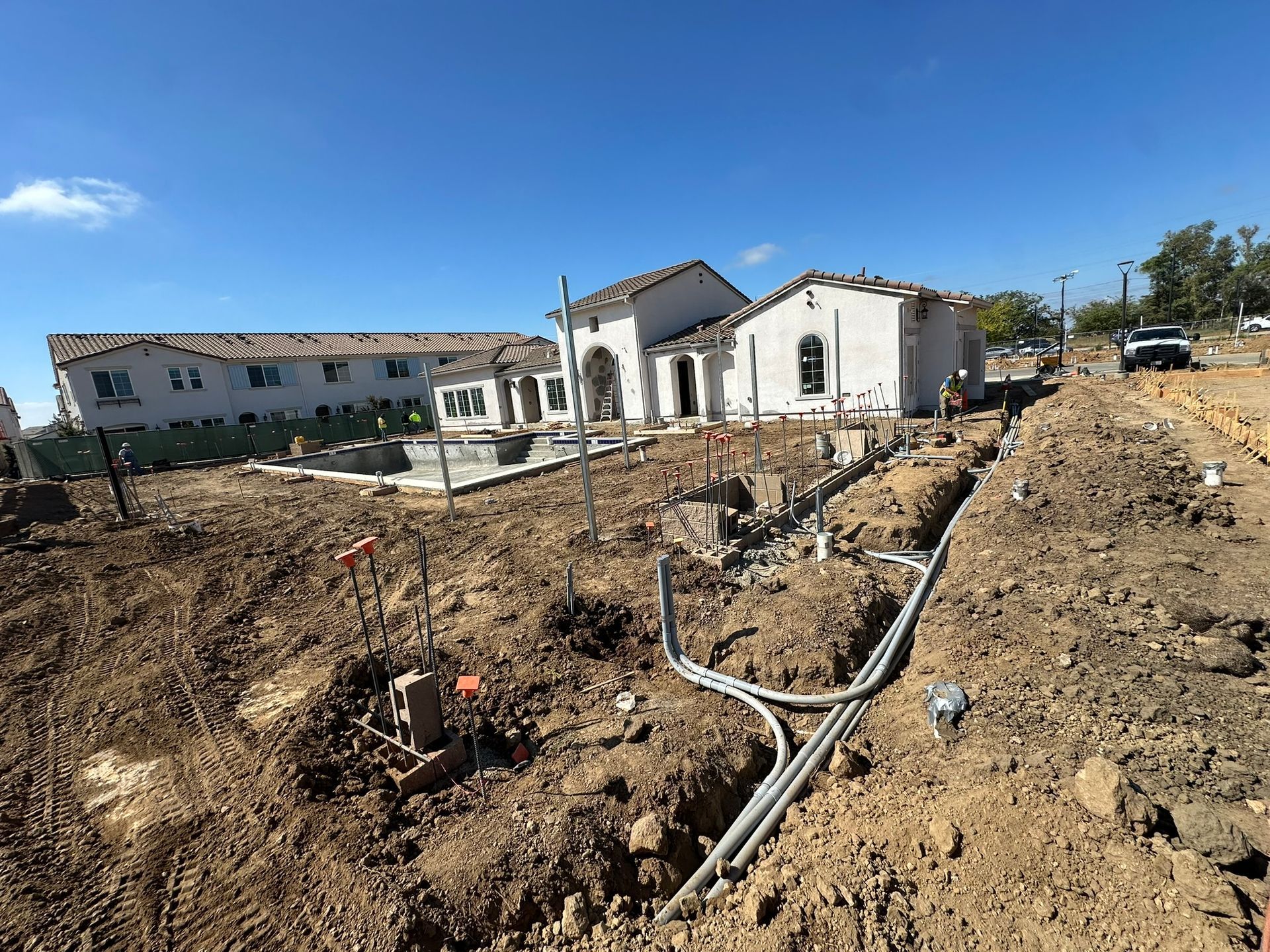 A construction site showing a new white house, a dirt lot, trenches, and electrical conduits under a clear blue sky.
