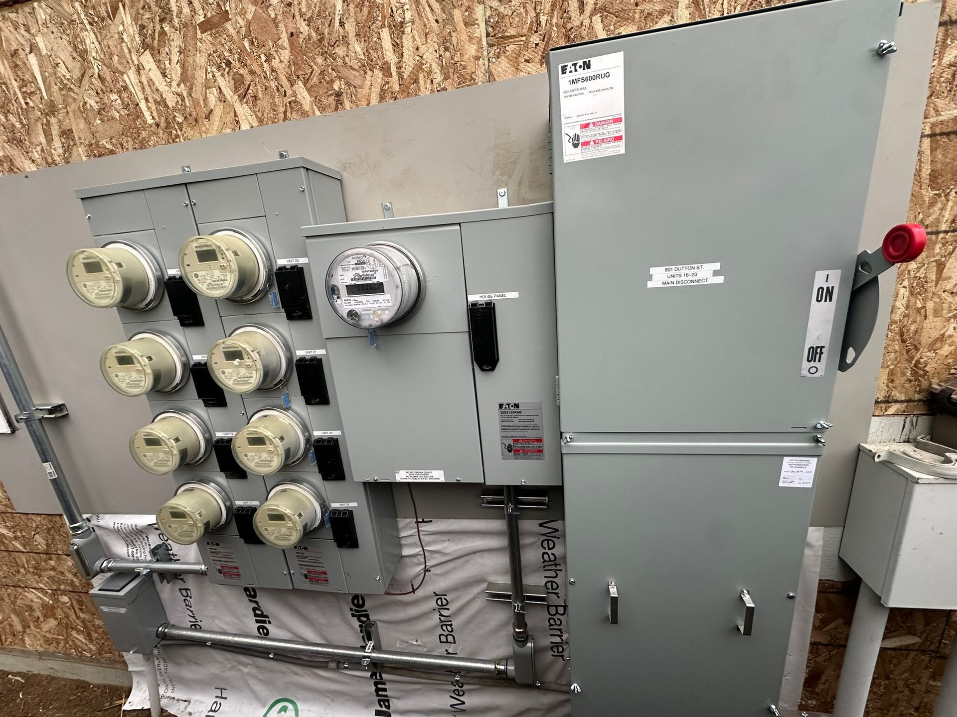 A gray electrical metering bank and large disconnect switch mounted on a wall with exposed wooden studs.