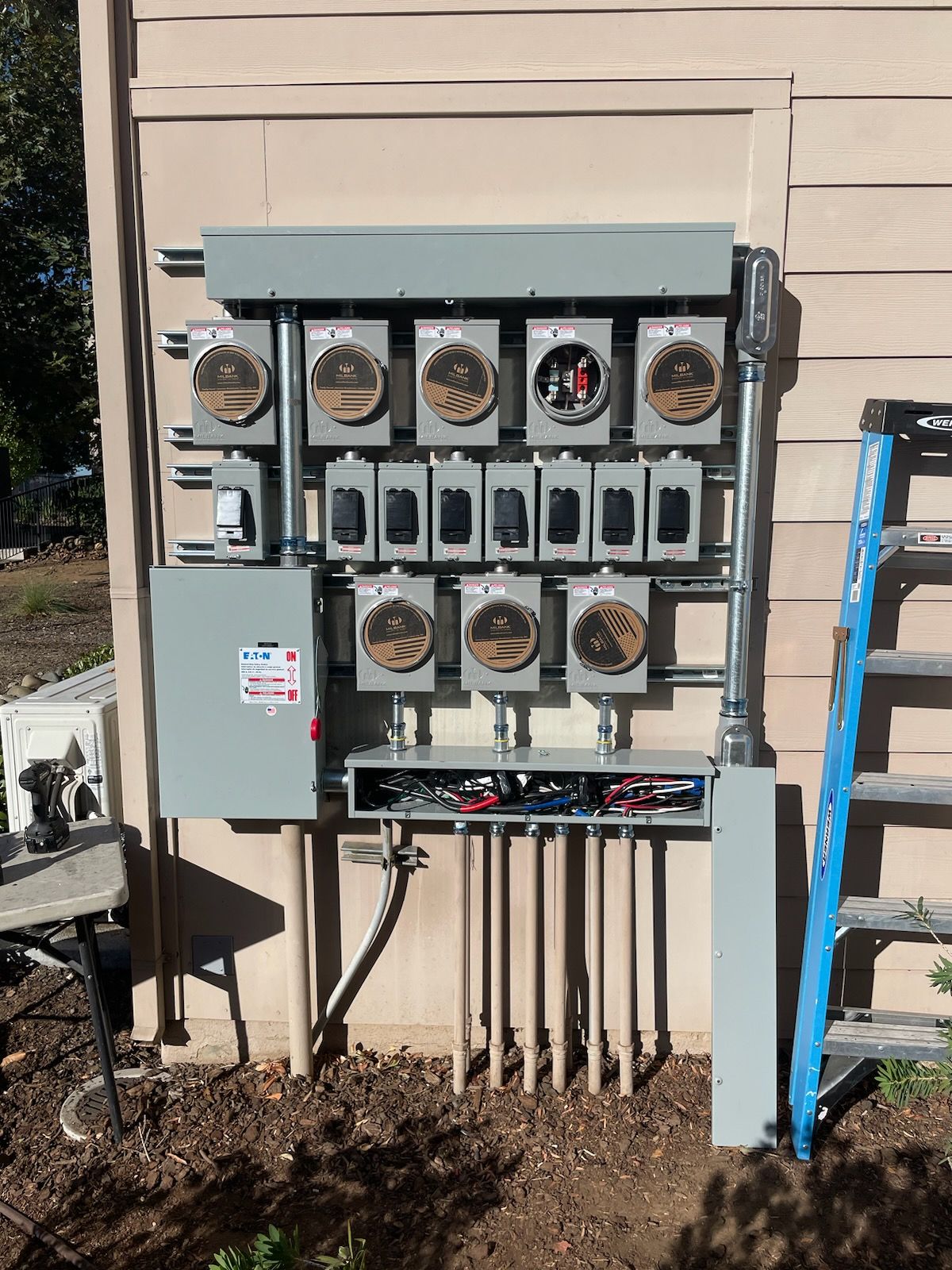 A grey electrical meter bank mounted on an exterior wall with multiple meters, disconnects, and conduit piping.