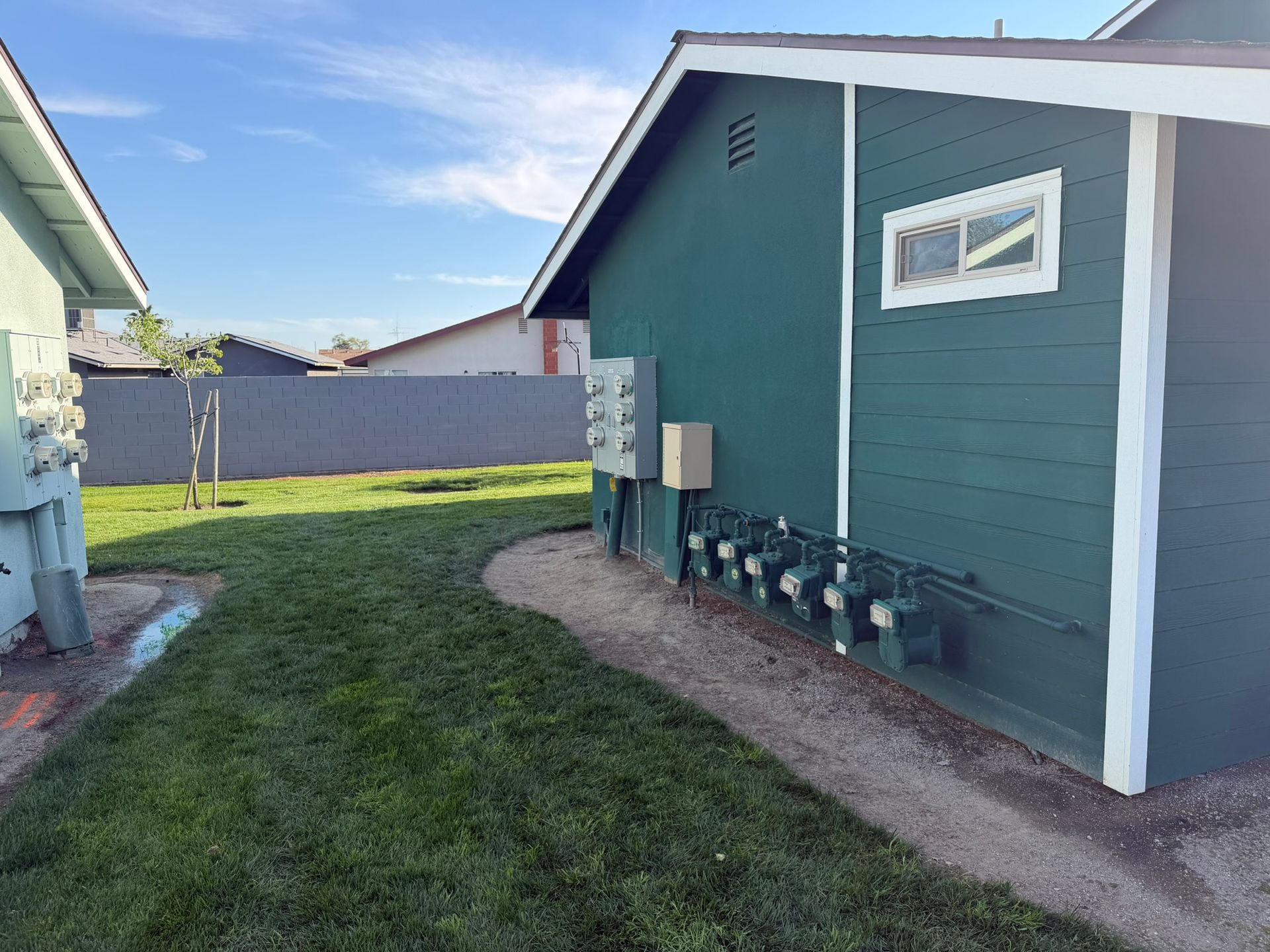 A dark green building exterior featuring multiple electrical meters and a window, situated next to a grassy yard.