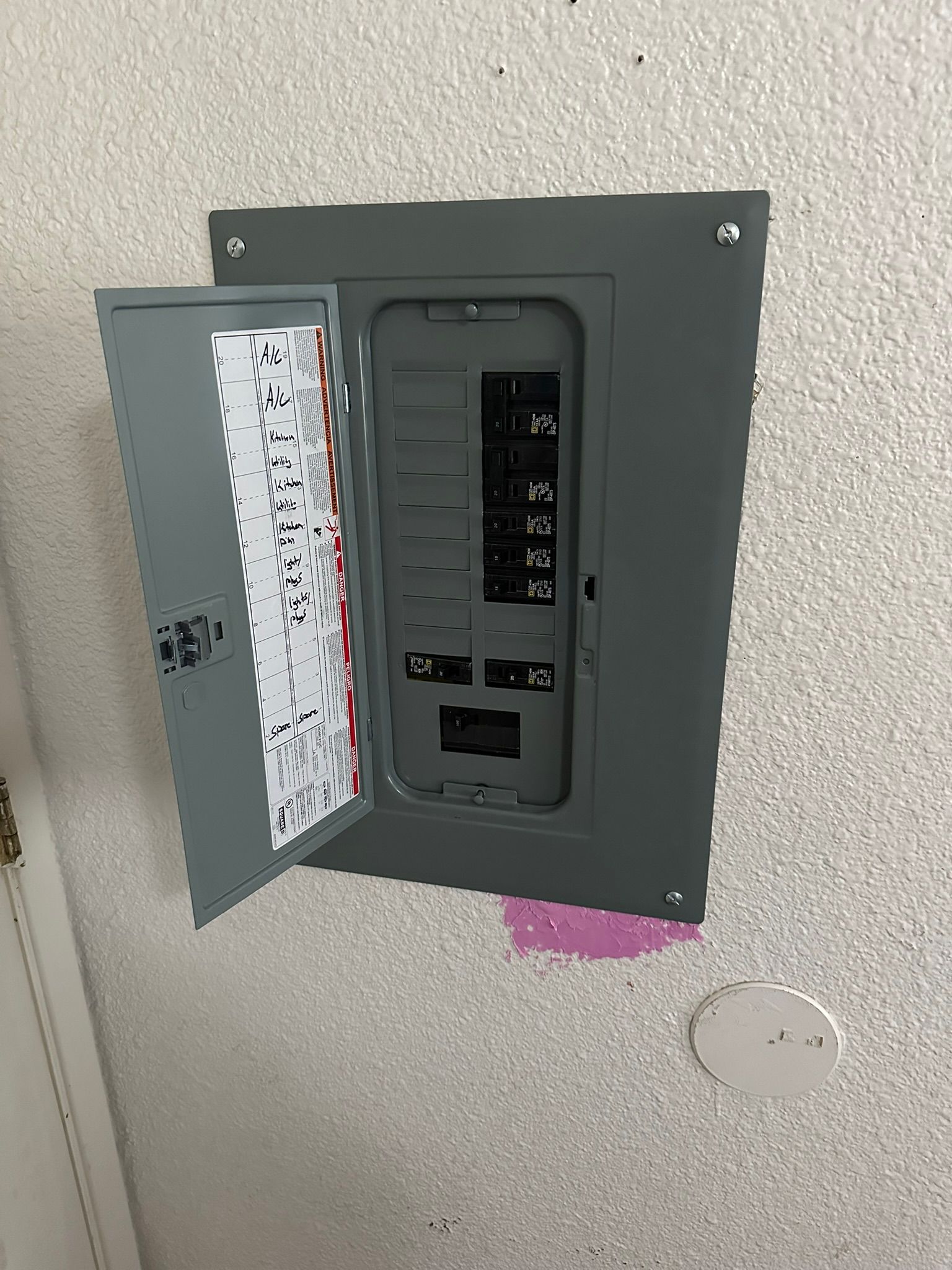 A gray residential electrical breaker panel mounted on a textured white wall, with its door open to reveal internal switches.
