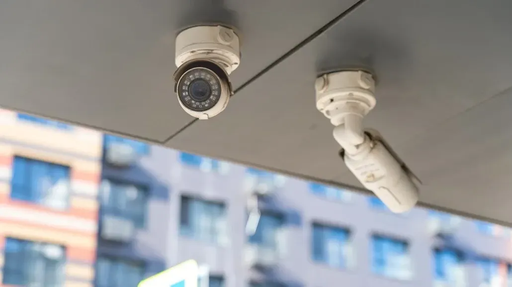 Two white security cameras mounted on the underside of a ceiling against a blurred building background.